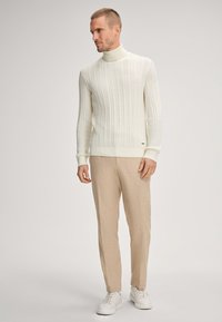 Witte kabelgebreide coltrui, beige broek en witte sneakers. Focus op het gestructureerde breipatroon en de getailleerde pasvorm.