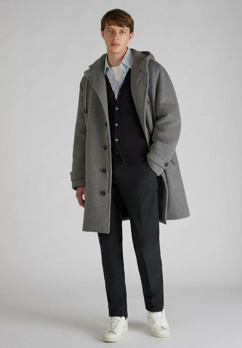 Cappotto di lana grigio con cappuccio, chiuso davanti con bottoni e tasche laterali, indossato sopra un cardigan nero e una camicia bianca, abbinato a pantaloni scuri e sneakers bianche.