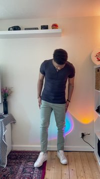 A person stands wearing a dark striped short-sleeve polo, light green pants, and white sneakers on a wooden floor.