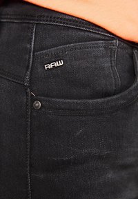 Jeans de mezclilla negra con una etiqueta del logo RAW visible, botón de metal y una textura consistente con la tela de mezclilla típica.