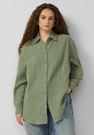 Kobieta o kręconych włosach, nosząca złote kolczyki-koła, luźną oliwkowozieloną koszulę na guziki oraz jasnoniebieskie dżinsy, stojąca na tle jednolitego tła.