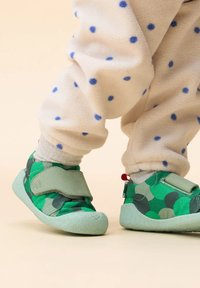 Scarpe verdi per bambini con un motivo a pois, realizzate in morbido tessuto con una chiusura in velcro. Suola di un verde chiaro con una texture. Indossate con pantaloni in pile a pois.