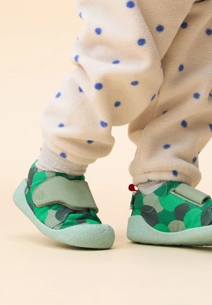 Groene kinderschoenen met een stippenpatroon, gemaakt van zacht stof met een klittenbandsluiting. Getextureerde lichtgroene zool. Draag met een fleece broek met stippen.