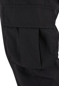 Gros plan d'une poche de pantalon cargo noire avec rabat et couture visible sur un tissu texturé.
