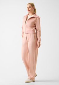Survêtement de sport rose pâle comprenant une veste zippée avec col et un pantalon assorti à jambes larges. Tissu lisse avec des détails de coutures discrètes.