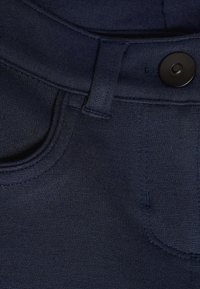 Detailný záber na tmavomodré nohavice zobrazujúci pás, pútko na opasok, gombík a švy na prednom vrecku.