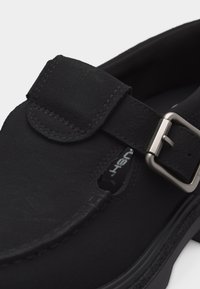 Μαύρο παπούτσι από σουέτ σε στυλ Mary Jane με ρυθμιζόμενο λουράκι και ασημένια αγκράφα, και ορατή ραφή σε λευκό φόντο.