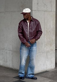 Veste en cuir bourgogne, chemise légère, jeans évasés bleu clair usés, chaussures noires, et une casquette blanche et bordeaux, avec des bagues aux mains.