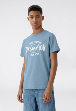 Jeune homme portant un t-shirt de sport bleu Champion avec texte blanc et jean bleu, debout devant un fond uni clair.