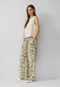 Romig mouwloos topje gecombineerd met losse, geprinte groene en beige broeken met grote bloemenontwerpen. Model draagt witte sandalen.