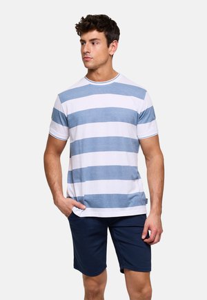 Joven de pie con una mano en el bolsillo, vistiendo una camisa de manga corta blanca con rayas azul claro y pantalones cortos azul marino, mirando hacia un lado.