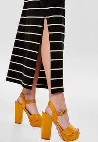 Gonna maxi a strisce nera e gialla con una fessura laterale, abbinata a sandali con tacco a piattaforma in suede giallo, caratterizzati da un cinturino a fibbia e un tacco spesso.