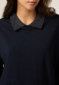 Top azul marino con un collar negro texturizado adornado con tachuelas metálicas, que destaca por su tejido suave y diseño clásico.