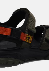 Brown and green sandal featuring a textured fabric upper, orange strap with a black buckle, and a rugged black outsole.