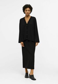 Donna con capelli corti e ricci, che indossa un cardigan nero a bottoni con scollo a V, una lunga gonna nera e scarpe mule a zeppa nere, in piedi su uno sfondo bianco.