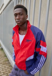 Crvena, plava i bijela zip-up vjetrovka s prugastim rukavima i logotipom. Tkanina izgleda lagano i teksturirano. Ima stojeći ovratnik.