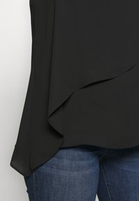 Svart flödande blus med böjd fåll, tillverkad av mjukt tyg och med lagerdesign, bärs över blå denimjeans.