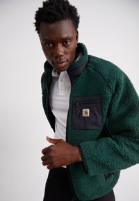 Grüne Fleecejacke mit schwarzen Akzenten, ausgestattet mit einer Reißverschlusstasche und hohem Kragen. Der strukturierte Stoff sorgt für ein gemütliches Aussehen.