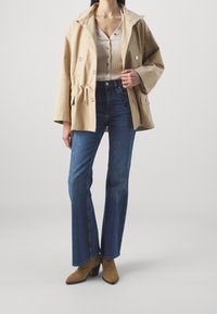 Chaqueta de algodón beige con cintura ajustable mediante cordón, cierre de botones y grandes bolsillos, combinada con jeans de campana azules y botas de tacón de tobillo en color beige.