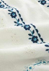 Tissu blanc avec broderie florale bleue texturée et délicats motifs floraux en découpe blanche sur un matériau léger.
