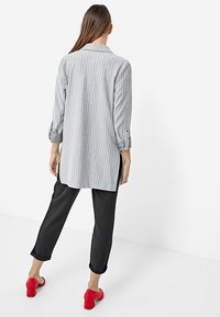 Vrouw met lang haar, gekleed in een grijs gestreepte lange blazer, zwarte cropped broek en rode hakken, met de rug naar de camera gericht op een witte achtergrond.