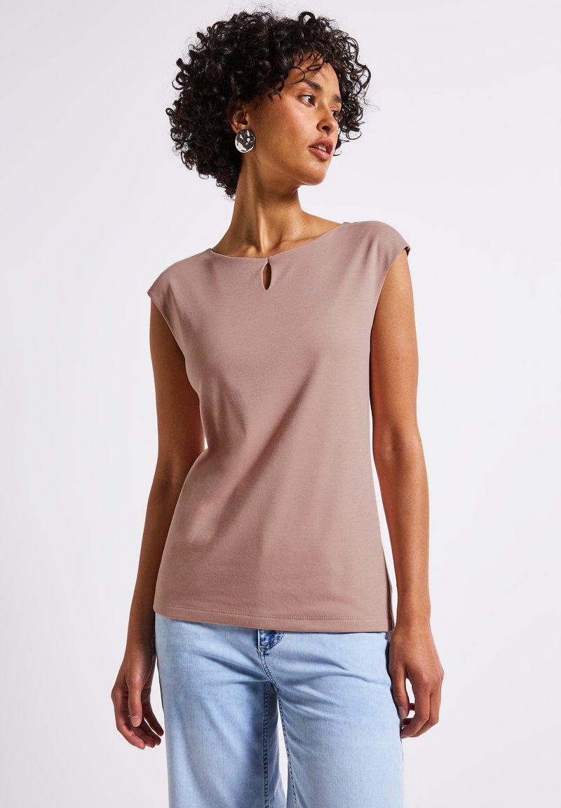 Beige přiléhavý top s krátkými zvonovými rukávy a malým detaily ve tvaru klíčové dírky na výstřihu. Hladká textura, spárované s světle modrými džíny.