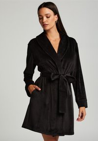 Svart sammetstol med sjalkrage, bältesmidja och långa ärmar. Har en mjuk textur och sidfickor. Enkelt och elegant design.