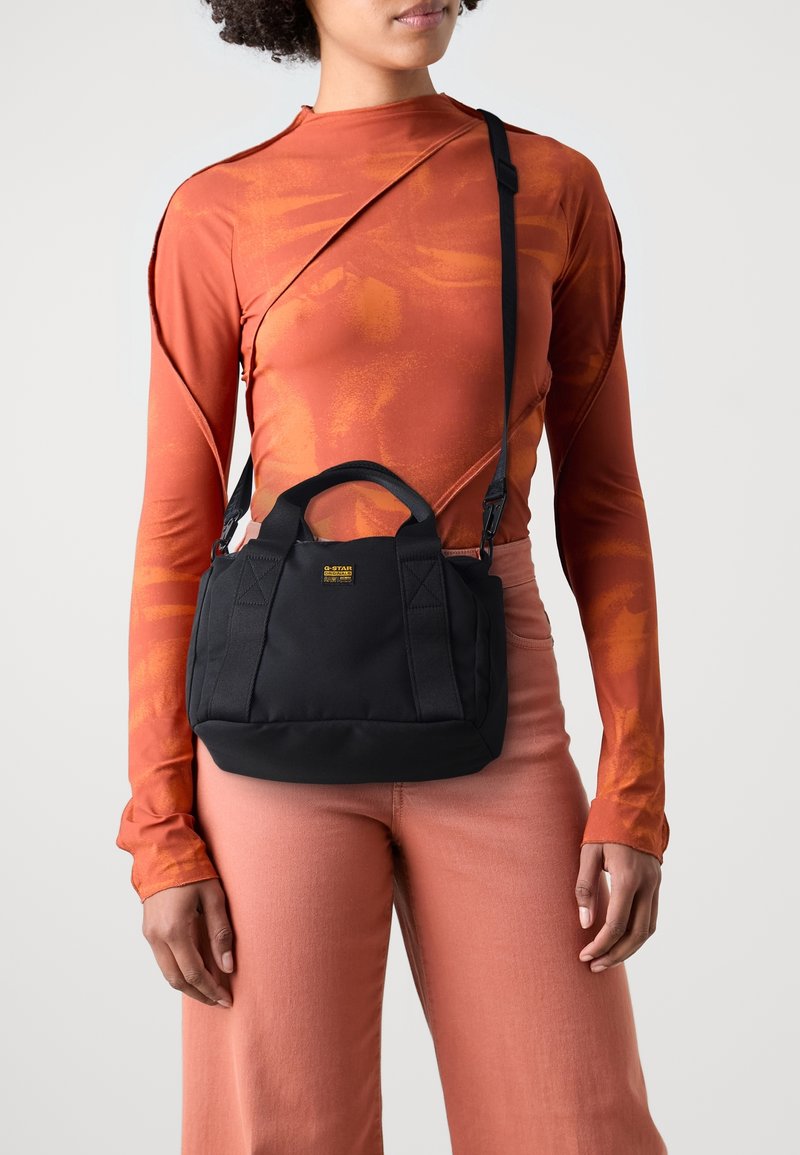 Personne portant un haut à manches longues orange brûlé et un pantalon rose, portant un petit sac bandoulière noir avec poignées et un écusson logo.