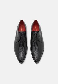 Un paio di scarpe eleganti in pelle nera lucida con dettagli wingtip e brogue, rivestimento interno rosso e lacci sul davanti, su sfondo bianco.