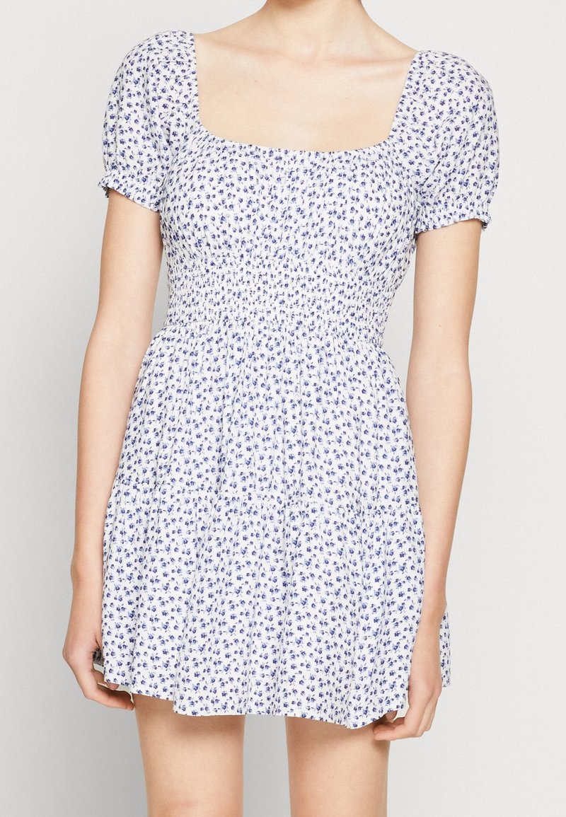 Květinové šaty se čtvercovým výstřihem, krátkými puff rukávy a nařaseným tělem. Bílé tkaniny s malými modrými květinami a rozšířenou sukní.