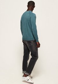 Maglione teal con collo a giro, realizzato in materiale morbido, abbinato a jeans grigio scuro e sneakers bianche. Presenta polsini e orlo a costine.