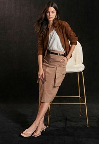 Brun ruskindsjakke over en hvid bluse, beige blyantskirt med cargolommer og tan hælede sandaler. Stol med gyldne ben i baggrunden.