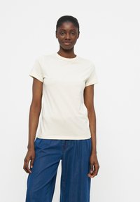 Γυναίκα που στέκεται στραμμένη προς τα εμπρός φορώντας απλό λευκό κοντομάνικο μπλουζάκι και άνετο μπλε παντελόνι μπροστά σε λευκό φόντο.