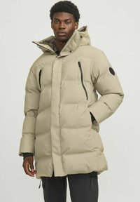 Beige puffer jacket with a high collar, featuring zippered pockets and elastic cuffs, paired with dark pants, standing against a light background.