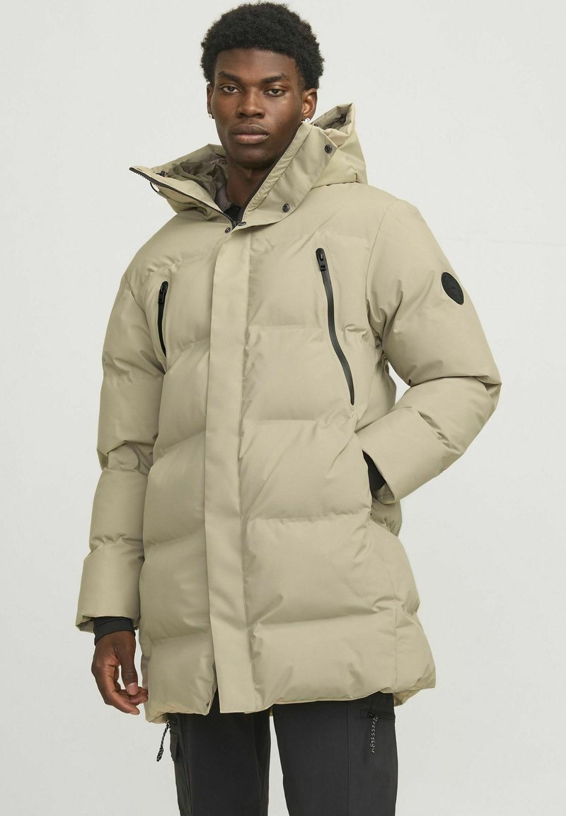 Beige puffer jacket with a high collar, featuring zippered pockets and elastic cuffs, paired with dark pants, standing against a light background.