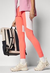 Coral leggings with white side panels, a fitted design, and a smooth texture. A beige and black backpack is held in the other hand.
