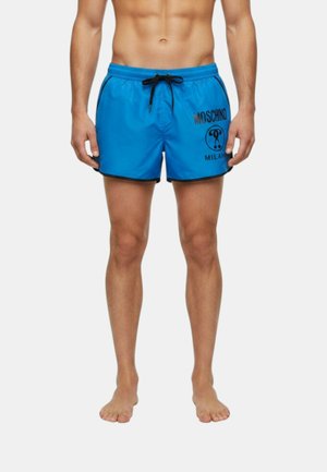 Uomo che indossa pantaloncini da bagno Moschino blu con coulisse e bordo neri, in piedi a piedi nudi su uno sfondo semplice.