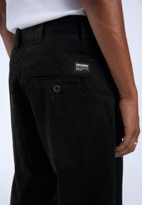 Los pantalones de mezclilla negra cuentan con un bolsillo trasero con cierre de botón y una etiqueta cuadrada que muestra "DRDENIM". La tela tiene una apariencia ligeramente texturizada.