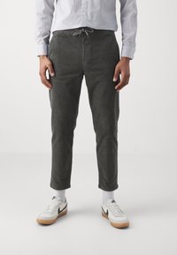 Pantalon en velours côtelé gris avec une taille à cordon, présentant des côtes verticales, une coupe fuselée et des détails de poches, porté avec des baskets blanches.