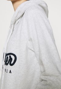 Sudadera gris claro con bolsillo frontal tipo canguro, capucha con cordón y logo bordado en azul marino en el pecho. Tejido suave y texturizado.