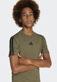 T-shirt a maniche corte verde oliva realizzato in tessuto morbido, con strisce diagonali nere sulle maniche e un piccolo logo nero sul petto.
