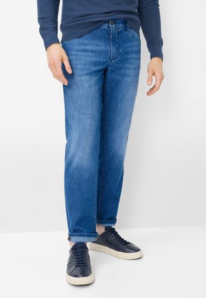 Uomo con jeans blu risvoltati, maglia a maniche lunghe blu navy e sneakers blu navy con suole bianche, in piedi contro uno sfondo bianco.