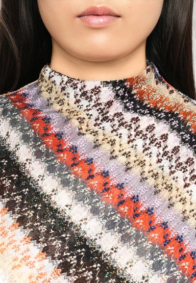 Nahaufnahme des unteren Gesichts und des Halses einer Frau, die einen mehrfarbigen, gemusterten Pullover mit hohem Kragen und strukturierten diagonalen Streifen trägt.