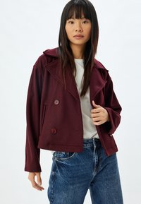giacca bordeaux doppiopetto con ampi revers, tasche laterali e chiusure con bottoni, realizzata in tessuto morbido, abbinata a jeans chiari.
