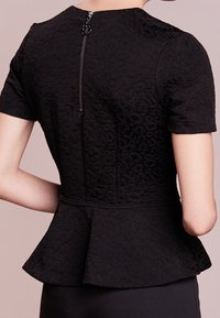 Musta tekstuuriga peplum-top, millel on lühikesed varrukad, peidetud tagumine tõmblukk ja kergelt reljeefne muster kangas.