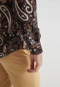 Chemise à manches longues en tissu noir avec un motif paisley en brun, blanc et or. Poignet boutoné avec des boutons blancs et des accents géométriques.