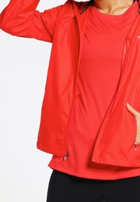 Chaqueta ligera roja con cremallera, sobre una camiseta roja ajustada a juego. Textura suave, mangas largas y bolsillos laterales visibles.