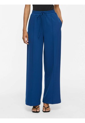 Personne portant un pantalon large bleu avec une taille élastique et un cordon, associé à des sandales noires et un top noir.
