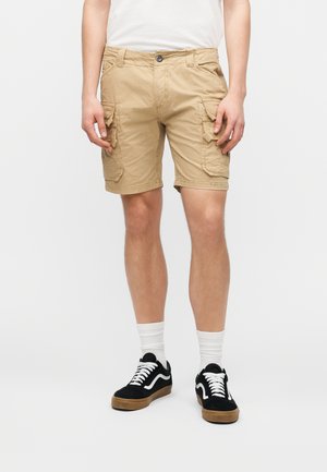 Persona que lleva pantalones cortos tipo cargo beige con múltiples bolsillos, calcetines blancos y zapatillas negras con cordones blancos, sobre un fondo liso.