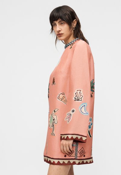 Γυναίκα με σκουρόχρωμα μαλλιά που φοράει ένα φαρδύ, ροζ φόρεμα με μακριά μανίκια, διακοσμημένο με πολύχρωμα αφηρημένα σχήματα και σχέδιο στο τελείωμα και τις μανσέτες.